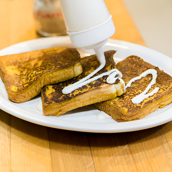 A white squeeze bottle dispenses white icing onto three slices of golden French toast on a white plate.